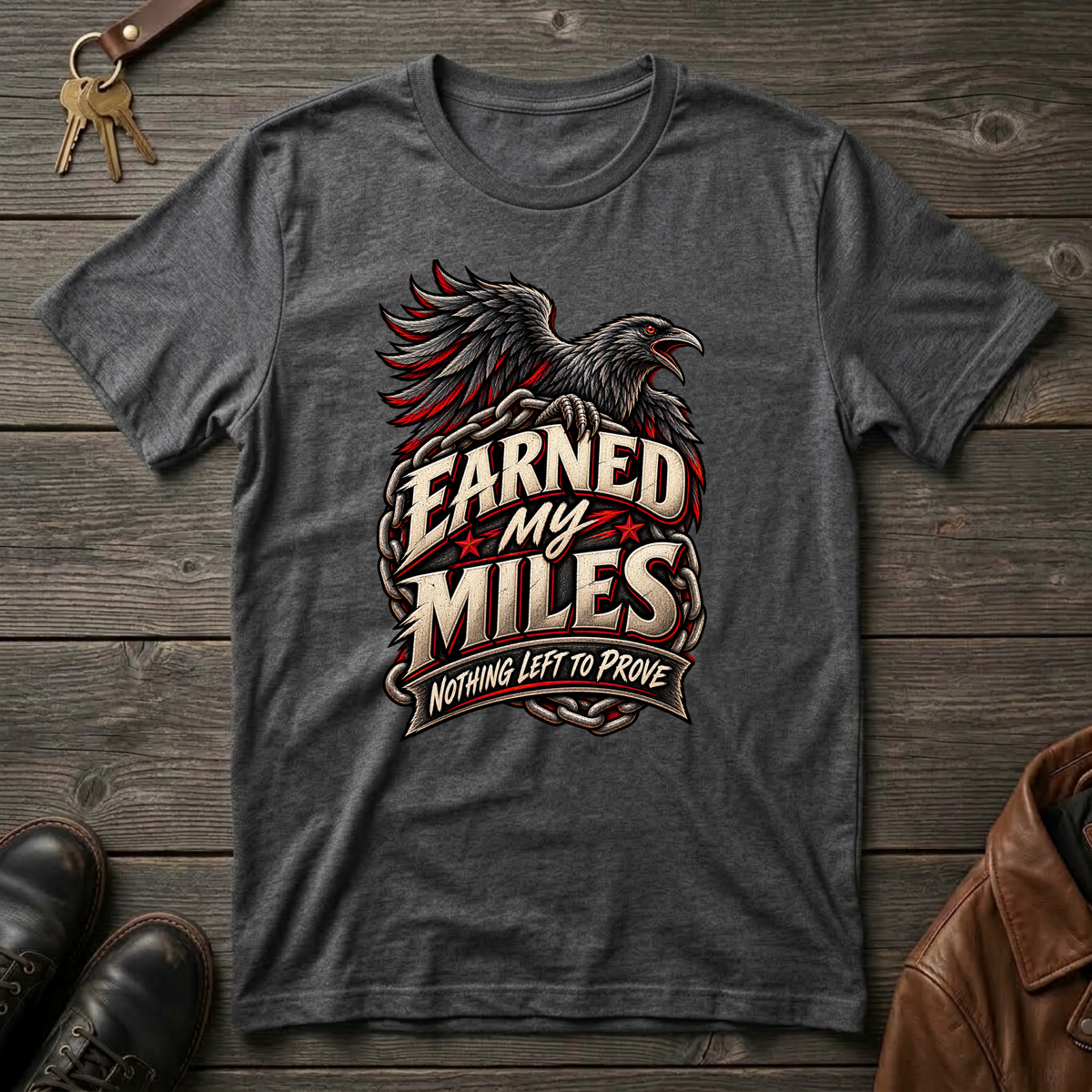 A dark heather gray graphic t-shirt featuring a raven and the text 'Earned My Miles Nothing Left to Prove', laid flat on a rustic dark wood background with keys, shoes, and a leather jacket as props.
