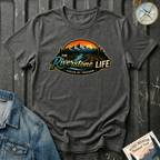 A dark heather graphic t-shirt laid flat on a dark textured background, styled with a blue denim jacket corner, a compass, and a Rocky Mountain National Park stamp. The t-shirt features a retro circular graphic of a river winding through mountains with the text 'The Riverstone Life Fueled by Freedom'.
