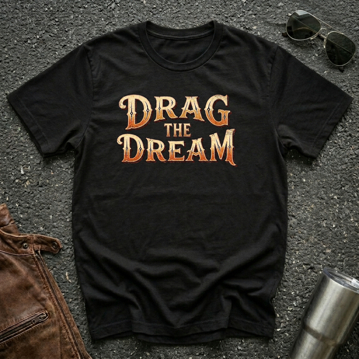 A black short-sleeve graphic T-shirt laid flat on a dark textured surface, with large orange and gold text reading “DRAG THE DREAM” across the chest. Sunglasses, a brown leather jacket, and a metal tumbler are placed around it as styling props.
