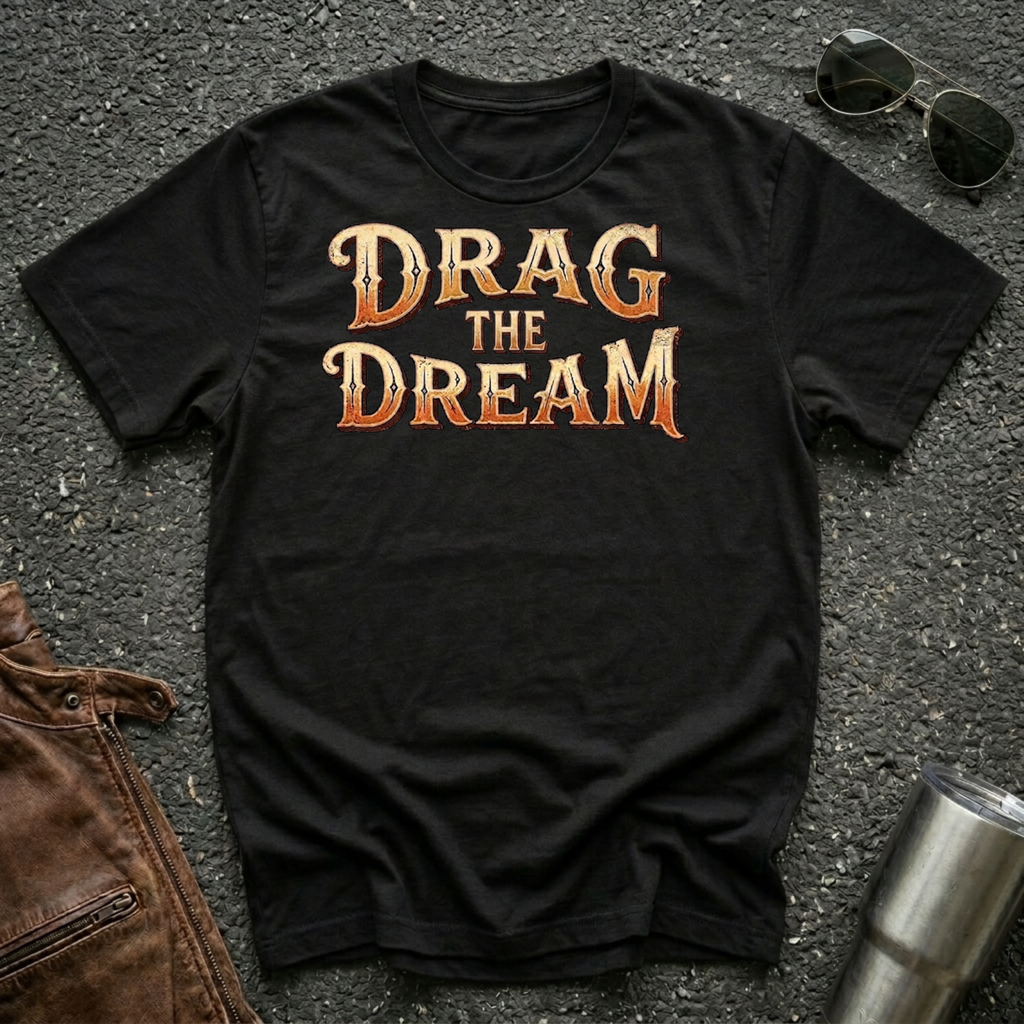 A black short-sleeve graphic T-shirt laid flat on a dark textured surface, with large orange and gold text reading “DRAG THE DREAM” across the chest. Sunglasses, a brown leather jacket, and a metal tumbler are placed around it as styling props.