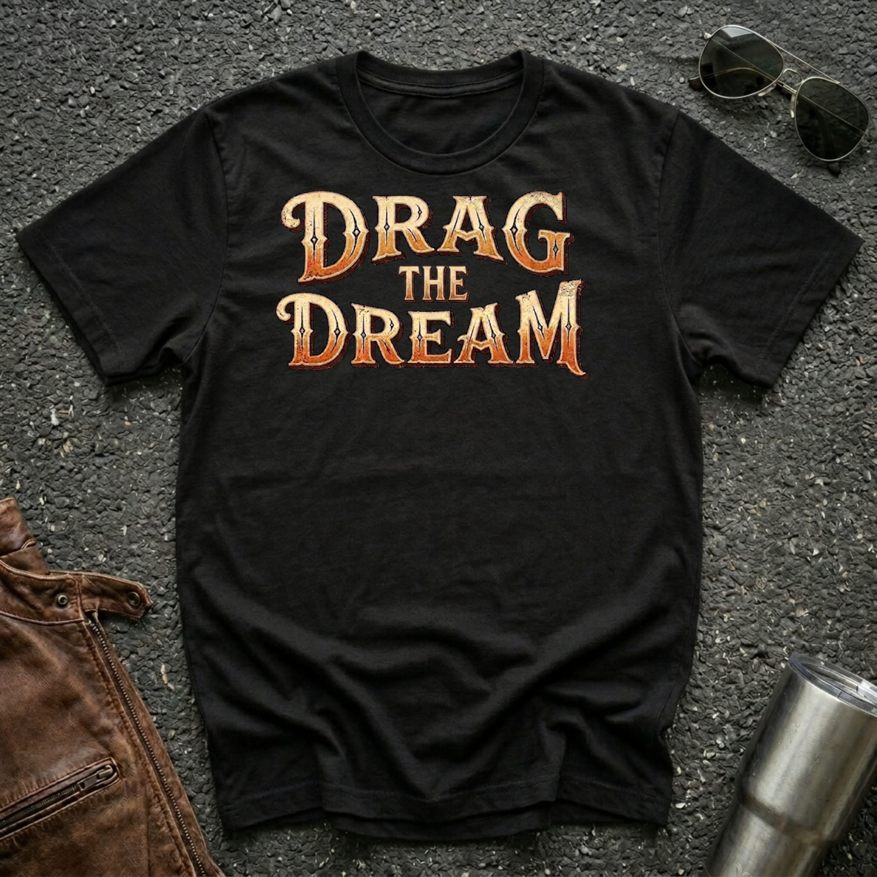 A black short-sleeve graphic T-shirt laid flat on a dark textured surface, with large orange and gold text reading “DRAG THE DREAM” across the chest. Sunglasses, a brown leather jacket, and a metal tumbler are placed around it as styling props.