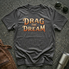 A dark heather short-sleeve graphic T-shirt laid flat on a dark textured surface, with large orange and gold text reading “DRAG THE DREAM” across the chest. Sunglasses, a brown leather jacket, and a metal tumbler are placed around it as styling props.