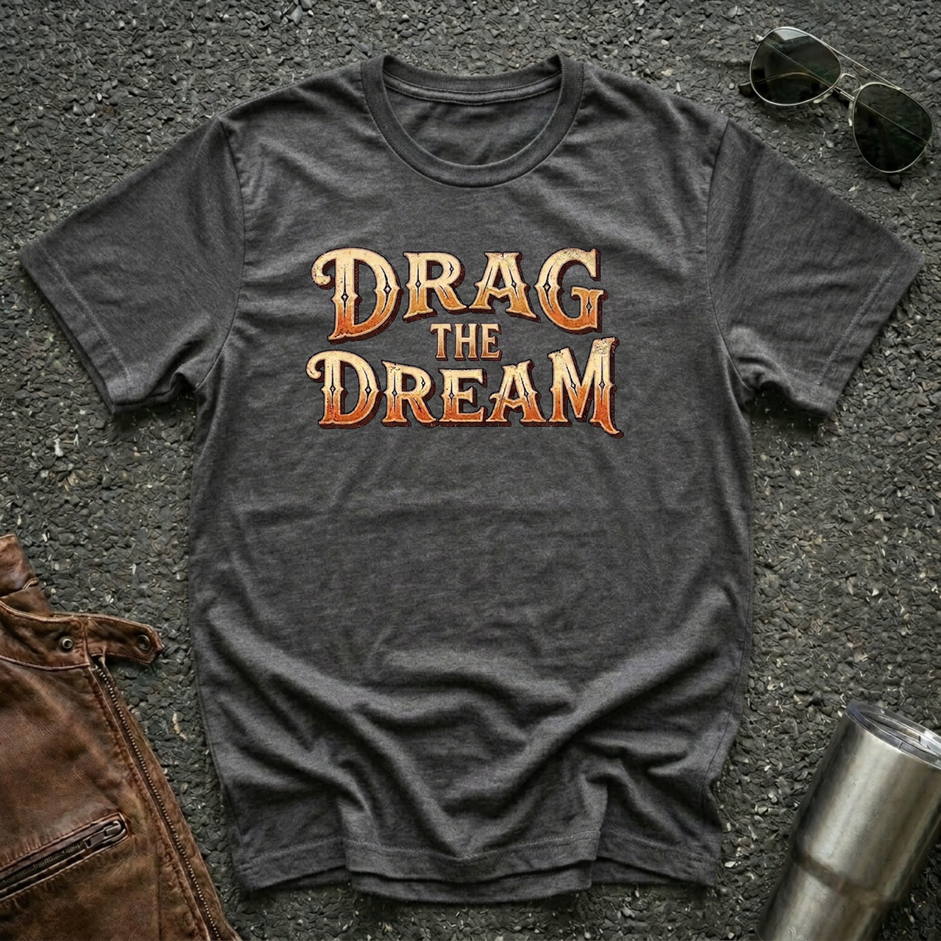 A dark heather short-sleeve graphic T-shirt laid flat on a dark textured surface, with large orange and gold text reading “DRAG THE DREAM” across the chest. Sunglasses, a brown leather jacket, and a metal tumbler are placed around it as styling props.
