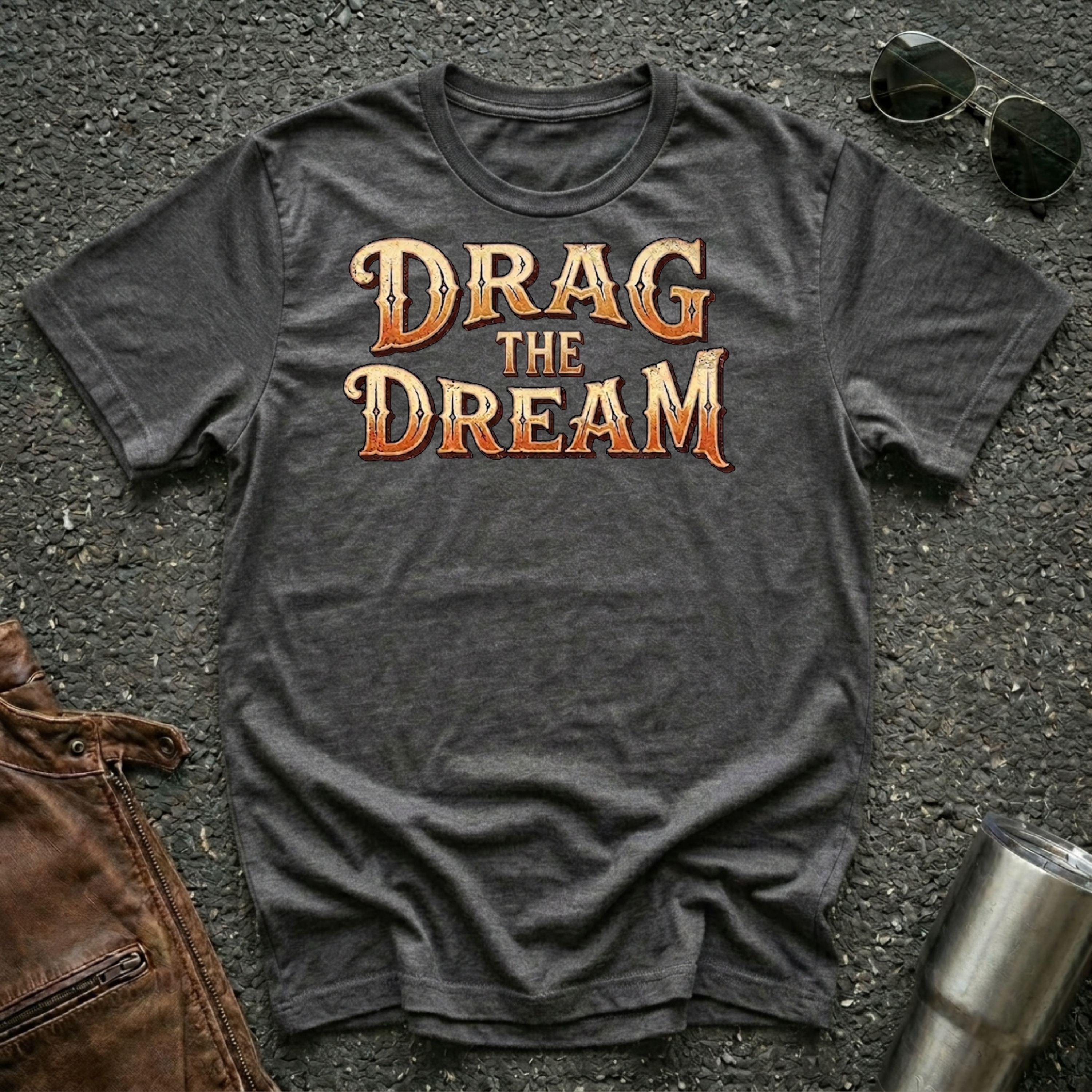 A dark heather gray graphic t-shirt laid flat on asphalt, featuring the phrase 'DRAG THE DREAM' in a stylized orange and gold font. The shirt is styled with sunglasses, a brown leather jacket, and a metal tumbler.