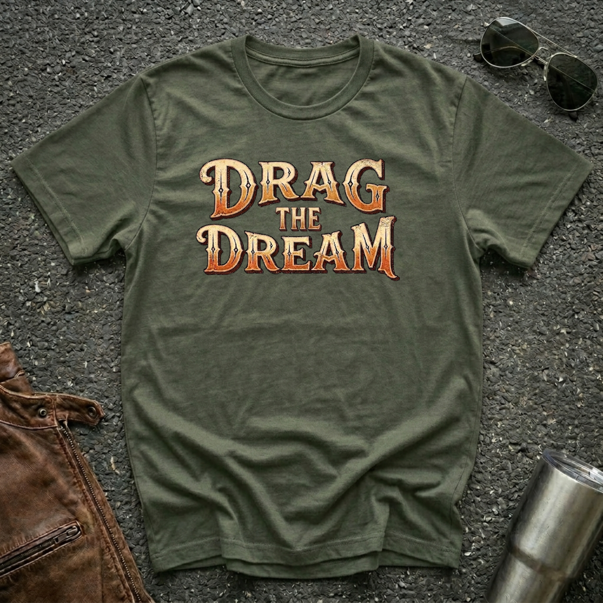 A military green short-sleeve graphic T-shirt laid flat on a dark textured surface, with large orange and gold text reading “DRAG THE DREAM” across the chest. Sunglasses, a brown leather jacket, and a metal tumbler are placed around it as styling props.