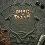 An olive green t-shirt with the phrase 'DRAG THE DREAM' printed in a stylized orange and brown font, laid flat on a dark asphalt surface with sunglasses, a brown leather jacket, and a metal tumbler nearby.