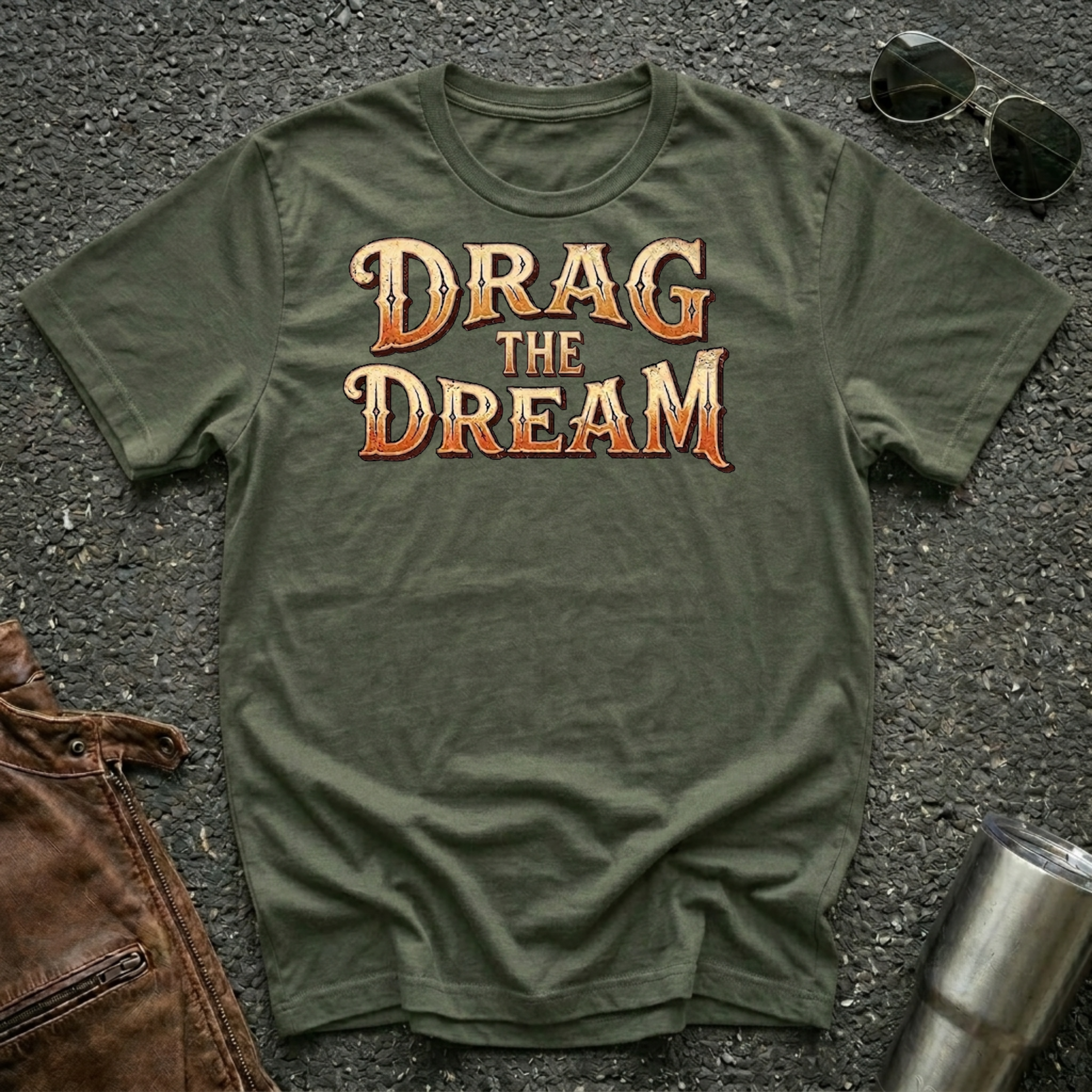 An olive green t-shirt with the phrase 'DRAG THE DREAM' printed in a stylized orange and brown font, laid flat on a dark asphalt surface with sunglasses, a brown leather jacket, and a metal tumbler nearby.