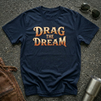 A navy short-sleeve graphic T-shirt laid flat on a dark textured surface, with large orange and gold text reading “DRAG THE DREAM” across the chest. Sunglasses, a brown leather jacket, and a metal tumbler are placed around it as styling props.