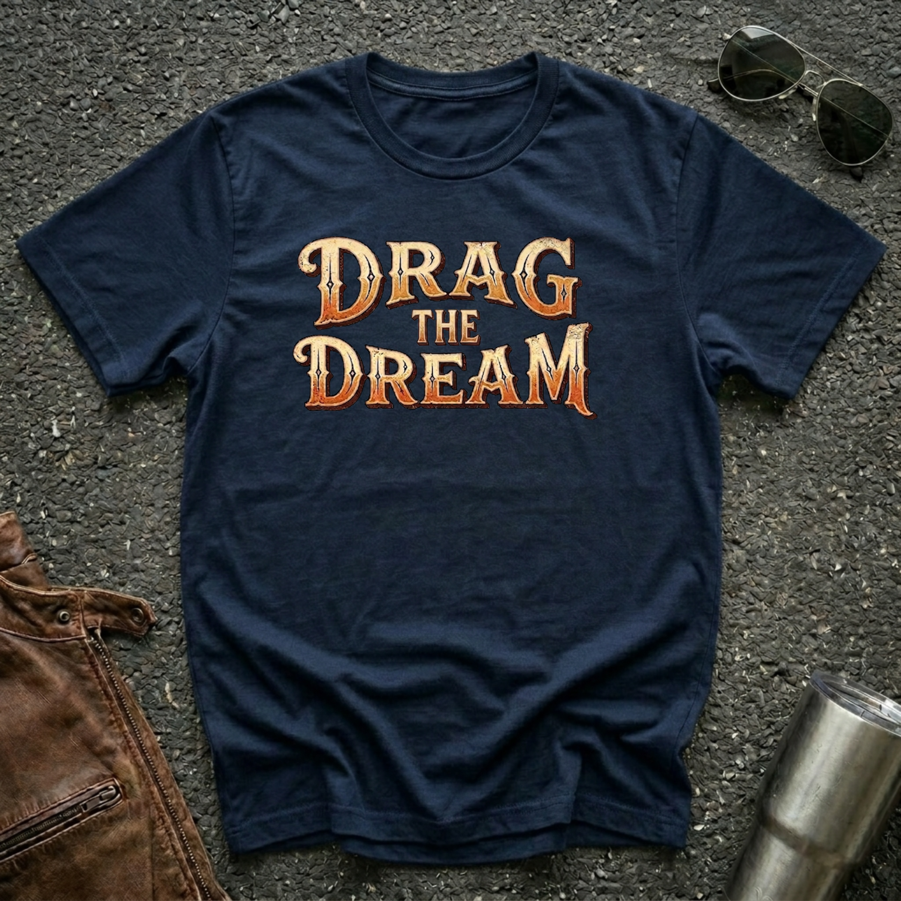 A navy short-sleeve graphic T-shirt laid flat on a dark textured surface, with large orange and gold text reading “DRAG THE DREAM” across the chest. Sunglasses, a brown leather jacket, and a metal tumbler are placed around it as styling props.