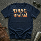 A navy blue graphic t-shirt with the phrase 'DRAG THE DREAM' printed in a textured orange and gold font, laid flat on a dark asphalt surface with sunglasses, a brown leather jacket, and a metal tumbler as styling props.