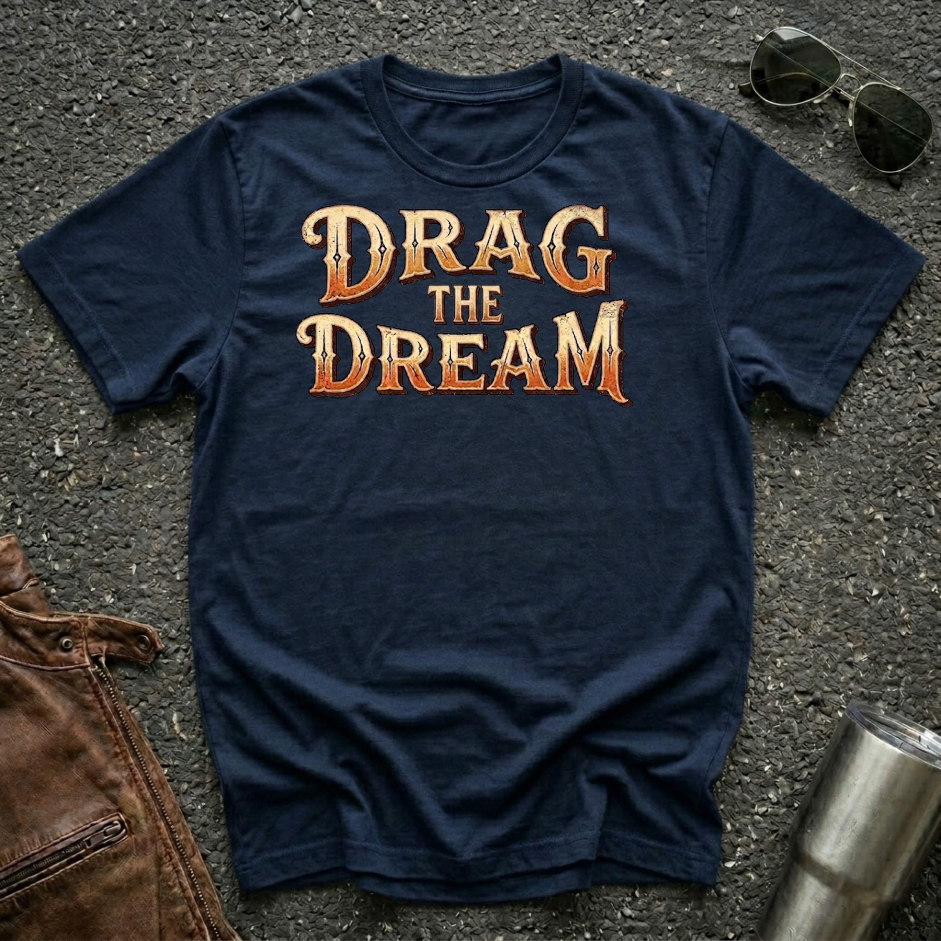 A navy blue graphic t-shirt with the phrase 'DRAG THE DREAM' printed in a textured orange and gold font, laid flat on a dark asphalt surface with sunglasses, a brown leather jacket, and a metal tumbler as styling props.
