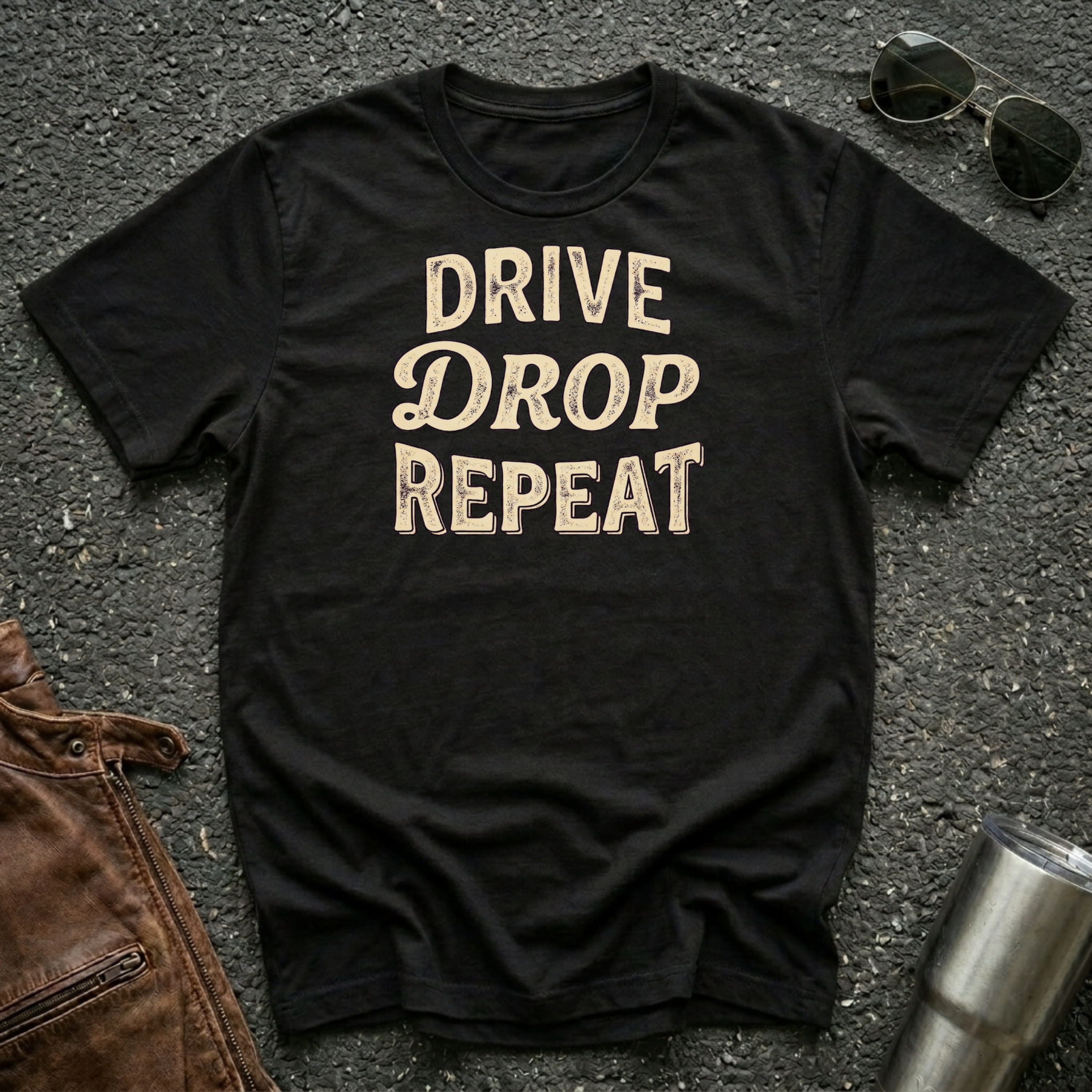 A black short-sleeve graphic T-shirt laid flat on a dark asphalt-like surface, with the phrase 'DRIVE DROP REPEAT' printed in large beige letters across the chest. Sunglasses, a brown leather jacket corner, and a metal tumbler are placed around it as styling props.