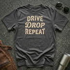 A dark heather gray graphic t-shirt with the words 'DRIVE DROP REPEAT' printed in a distressed beige font, laid flat on a gray asphalt surface with sunglasses, a brown leather jacket, and a stainless steel tumbler as props.