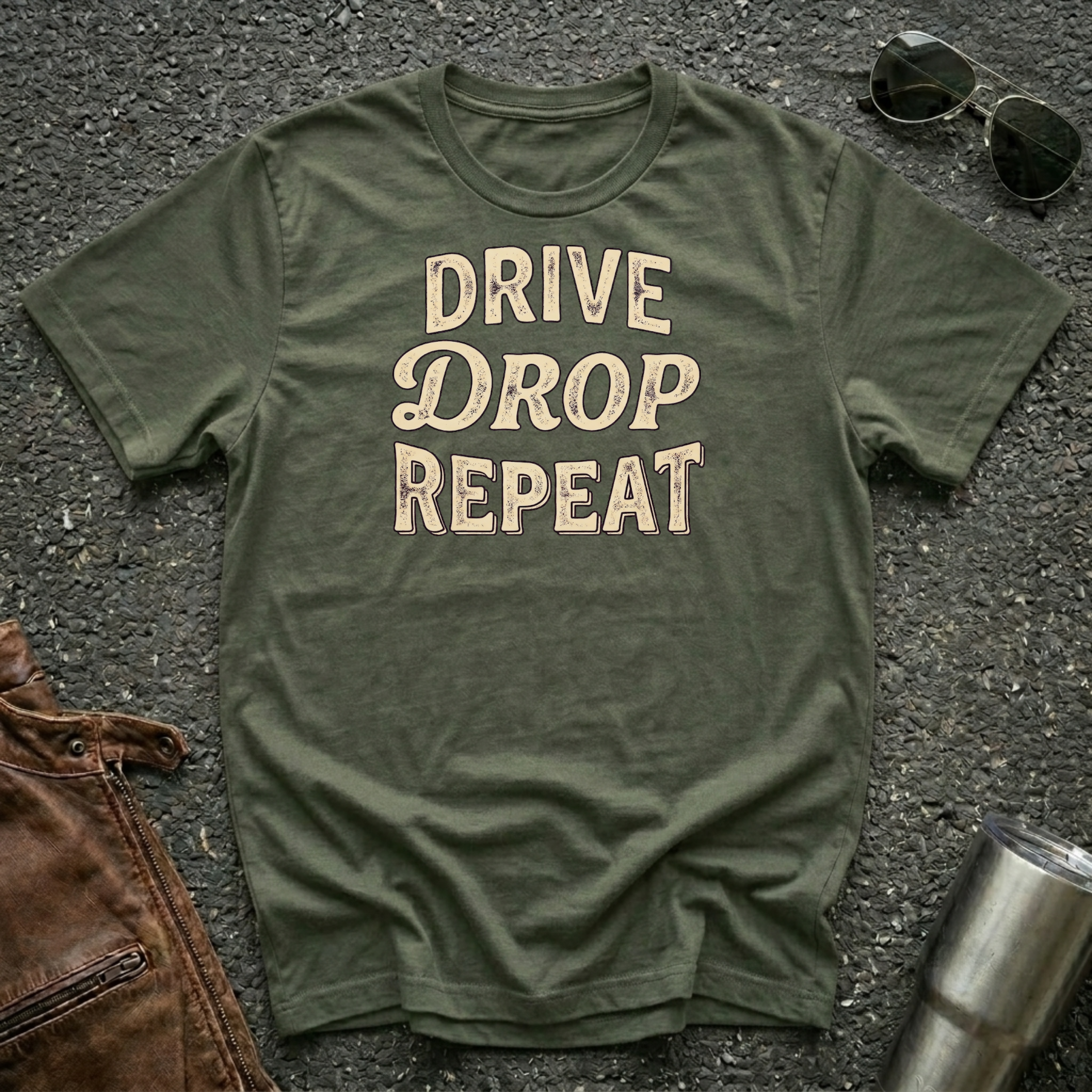 An olive green crewneck t-shirt with the text 'DRIVE DROP REPEAT' printed in a beige distressed font, laid flat on a gray asphalt surface with sunglasses, a brown leather jacket, and a metal tumbler as props.