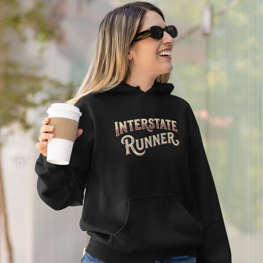 A woman wearing a black hoodie with 'INTERSTATE RUNNER' printed in brown and beige lettering, holding a coffee cup outdoors.