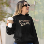 A woman wearing a black hoodie with 'INTERSTATE RUNNER' printed in brown and beige lettering, holding a coffee cup outdoors.