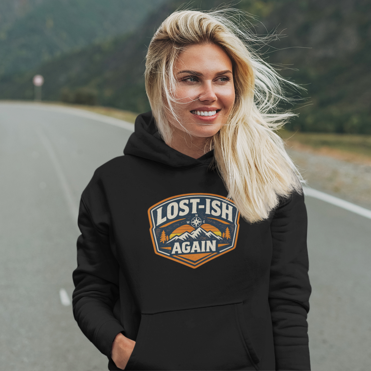 A smiling blonde woman stands on a paved road with mountains in the background, wearing a black hoodie with a large shield-style chest graphic that reads “LOST-ISH AGAIN” in white, blue, and orange.