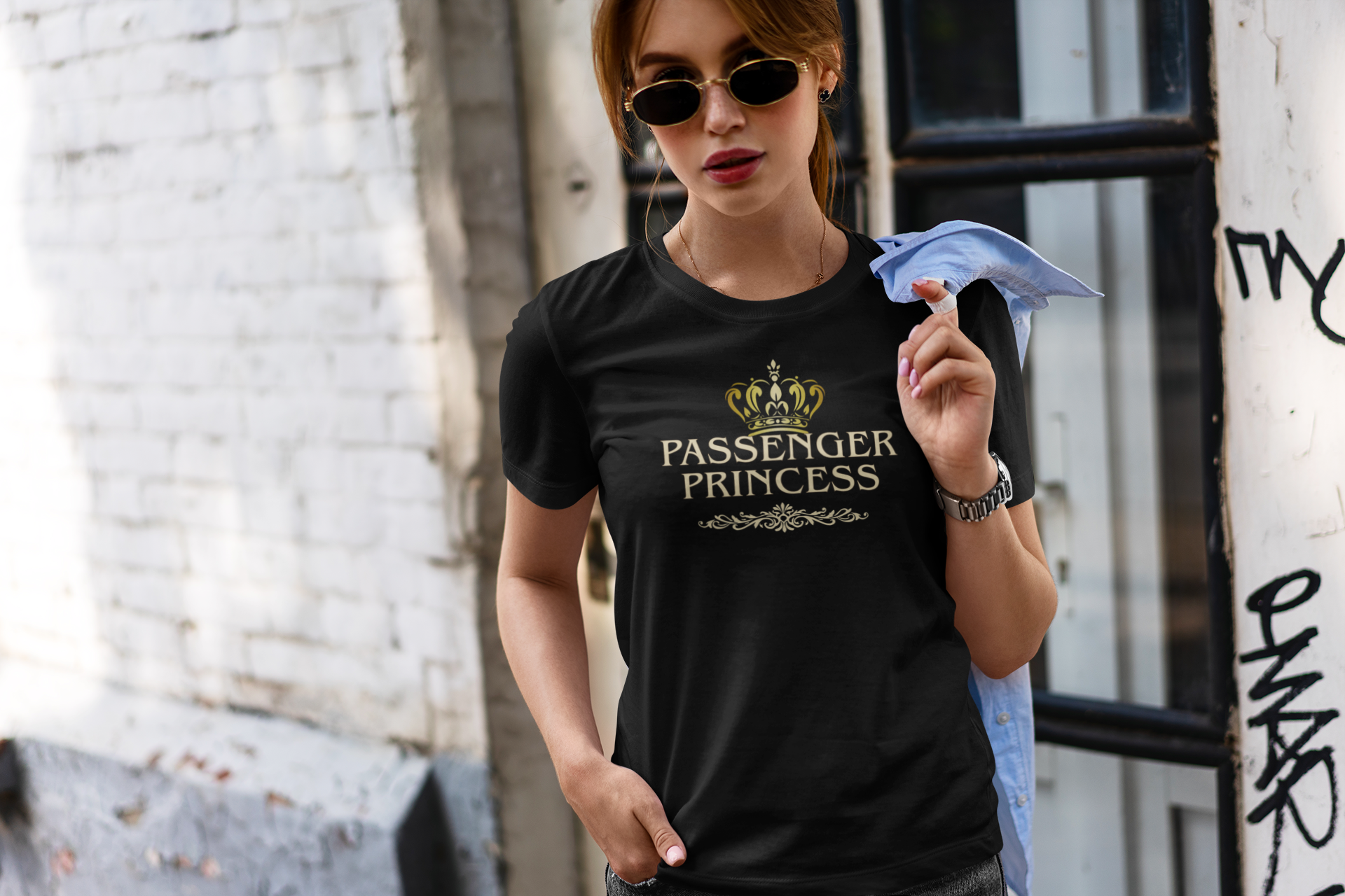 A woman wearing a black graphic t-shirt with a gold crown and 'PASSENGER PRINCESS' text, standing in an urban alleyway.