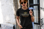 A woman wearing a black graphic t-shirt with a gold crown and 'PASSENGER PRINCESS NAVIGATION EXPERT' text, holding a light blue shirt in an urban alleyway.