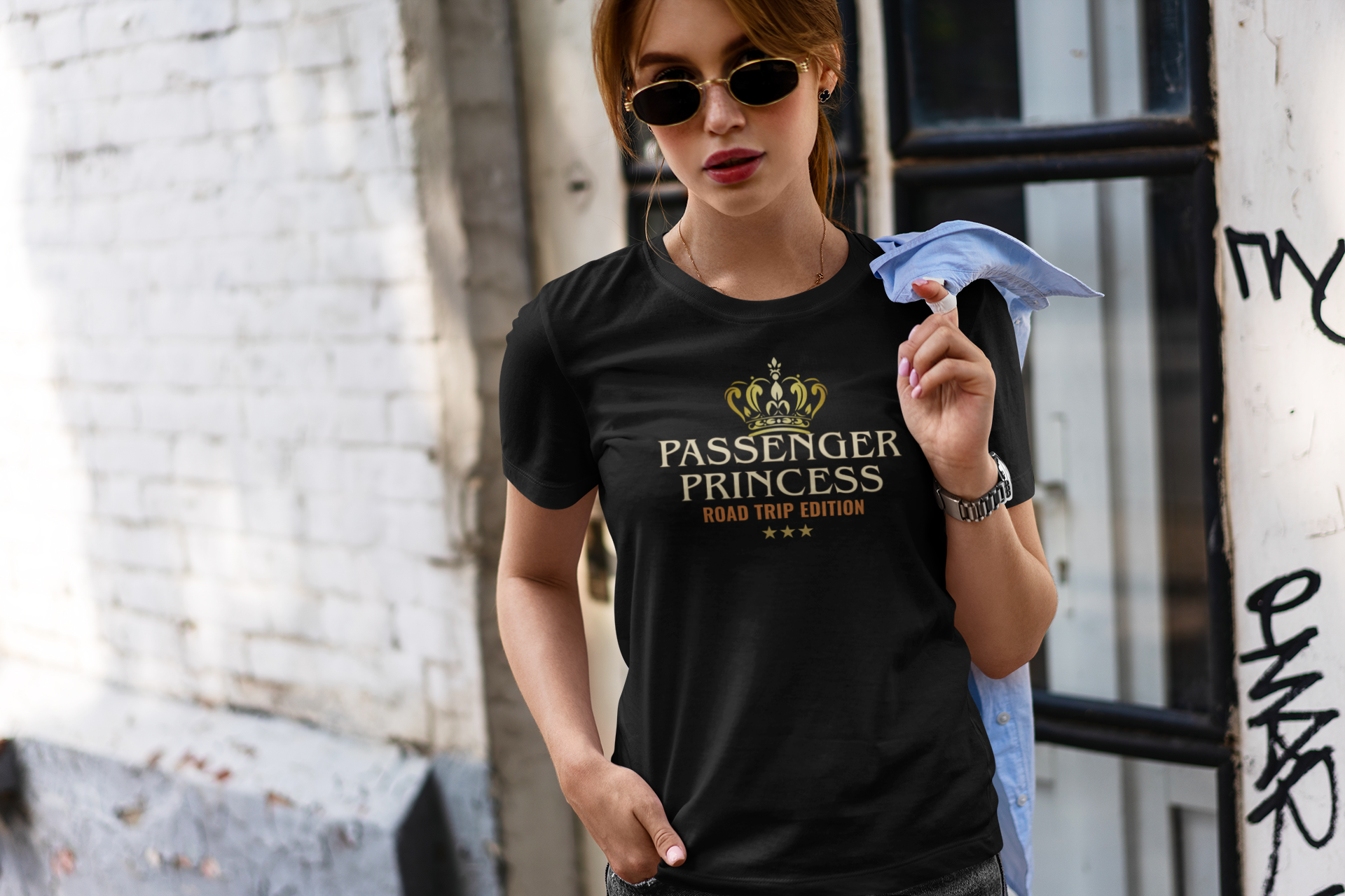 A woman wearing a black graphic t-shirt with a gold crown and 'PASSENGER PRINCESS ROAD TRIP EDITION' text, standing in an urban alleyway.