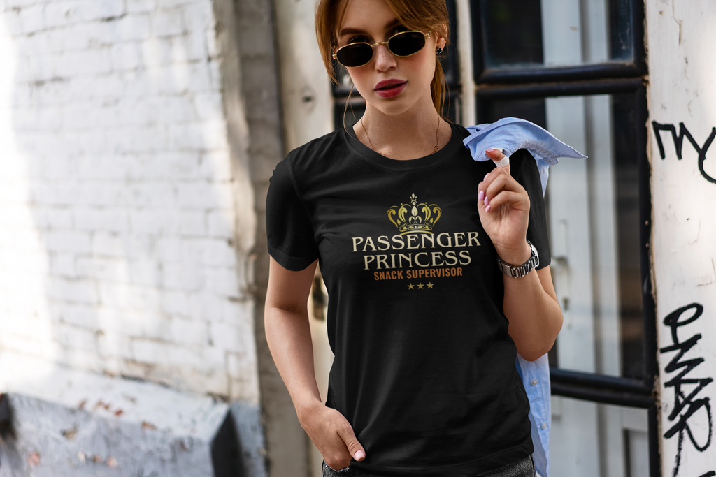 A woman wearing a black graphic t-shirt with a gold crown and the text 'PASSENGER PRINCESS SNACK SUPERVISOR' stands in an urban setting.