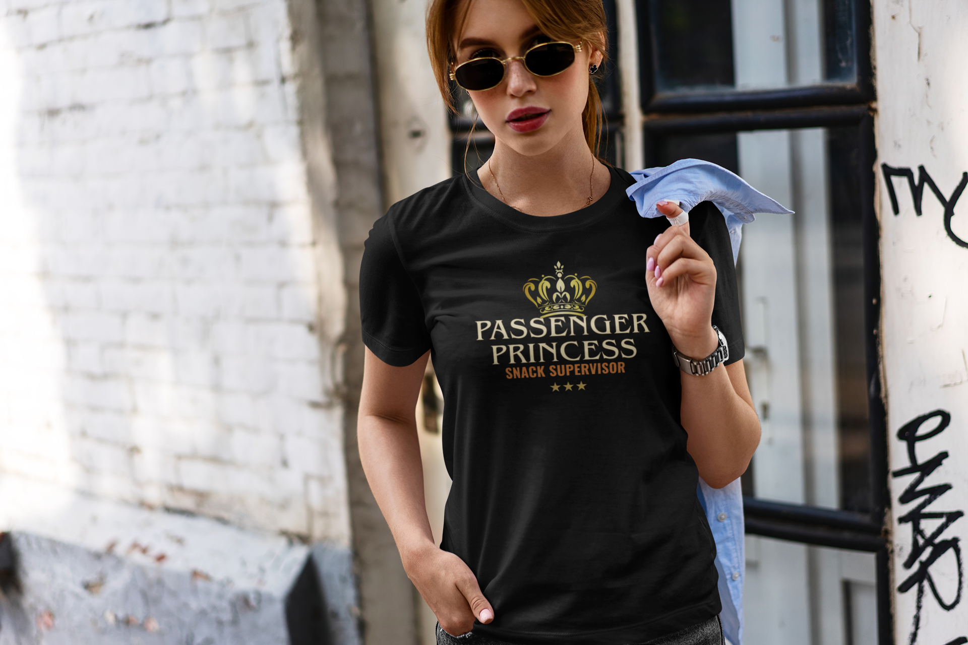 A woman wearing a black graphic t-shirt with a gold crown and the text 'PASSENGER PRINCESS SNACK SUPERVISOR' stands in an urban setting.