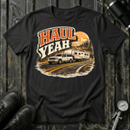 Haul Yeah RV Transport T-shirt by The Riverstone Life on a black Gildan Softstyle 64000 featuring a pickup truck towing a fifth-wheel RV.