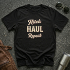 A black graphic t-shirt with the words 'Hitch HAUL Repeat' printed in a distressed cream and maroon font, laid flat on a dark asphalt surface with sunglasses, brown leather jacket, and a metal tumbler nearby.