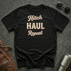 A black graphic t-shirt with the words 'Hitch HAUL Repeat' printed in a distressed cream and maroon font, laid flat on a dark asphalt surface with sunglasses, brown leather jacket, and a metal tumbler nearby.