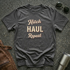 A dark heather graphic t-shirt with the words 'Hitch HAUL Repeat' printed in a distressed cream and maroon font, laid flat on a dark asphalt surface with sunglasses, brown leather jacket, and a metal tumbler nearby.