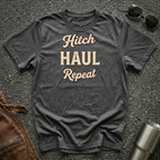 A dark heather gray graphic t-shirt laid flat on asphalt, featuring the words 'Hitch HAUL Repeat' in a cream and maroon retro font. Styled flat-lay props include sunglasses, a brown leather jacket, and a metal tumbler.