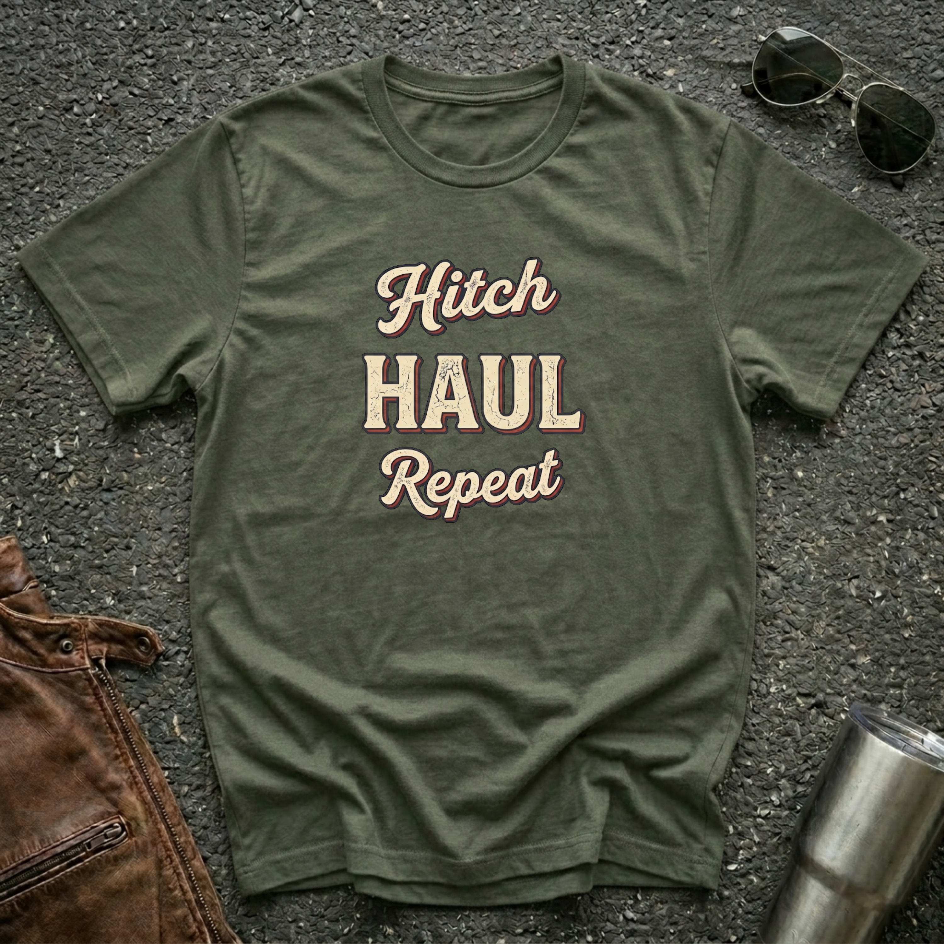 A military green graphic t-shirt with the words 'Hitch HAUL Repeat' printed in a distressed cream and maroon font, laid flat on a dark asphalt surface with sunglasses, brown leather jacket, and a metal tumbler nearby.