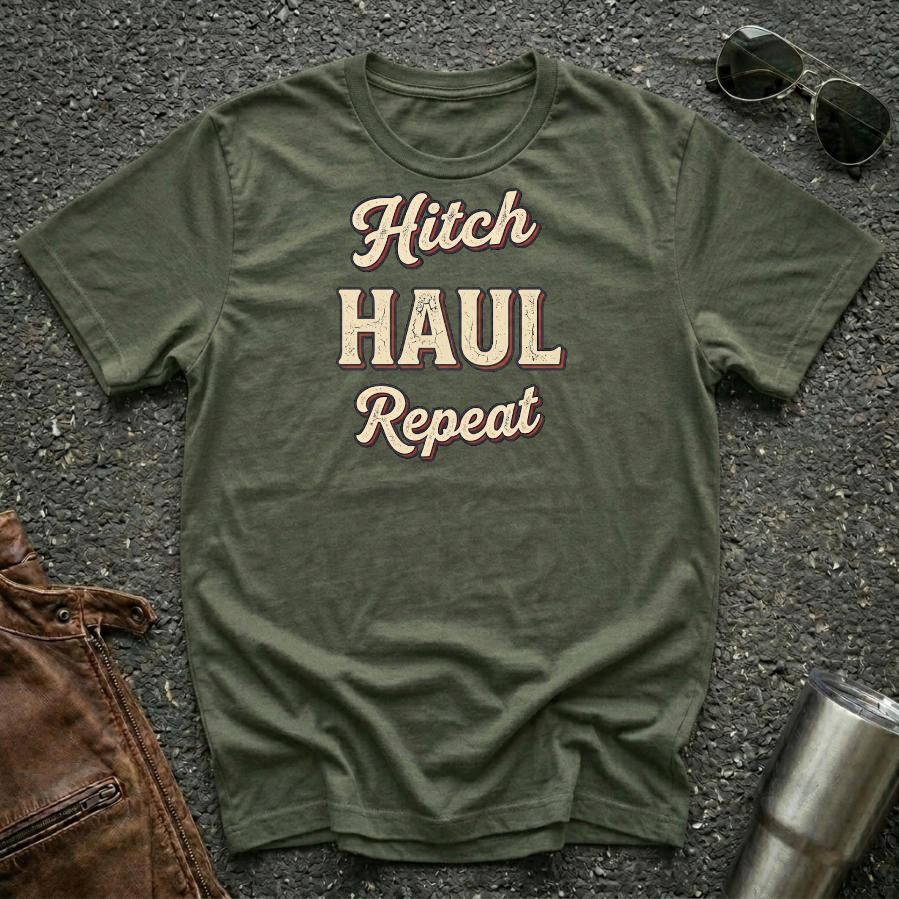 An olive green graphic t-shirt laid flat on a dark asphalt surface, featuring the words 'Hitch HAUL Repeat' in a beige and maroon retro font. Styled flat-lay props include sunglasses at the top right, a brown leather jacket at the bottom left, and a silver tumbler at the bottom right.