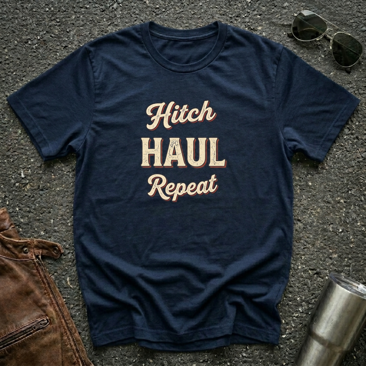 A navy graphic t-shirt with the words 'Hitch HAUL Repeat' printed in a distressed cream and maroon font, laid flat on a dark asphalt surface with sunglasses, brown leather jacket, and a metal tumbler nearby.