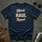 A navy blue graphic t-shirt with the words 'Hitch HAUL Repeat' printed in a beige and maroon retro font, laid flat on a gray asphalt surface with sunglasses, a brown leather jacket, and a metal tumbler nearby.