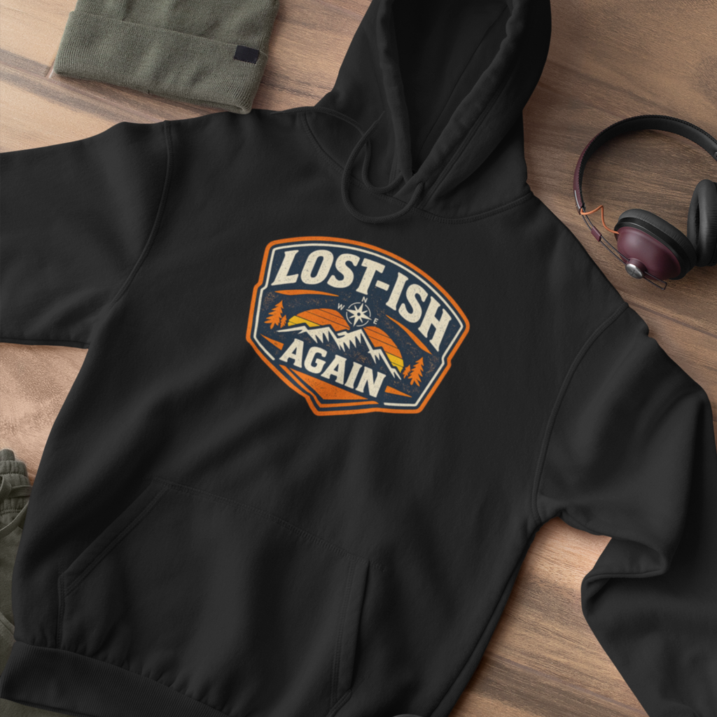 A black pullover hoodie laid flat on a wood surface, featuring a large chest graphic that reads “LOST-ISH AGAIN” with mountains, trees, and an orange sun. A folded olive green garment, headphones, and a small shoe edge are partially visible around it as styling props.