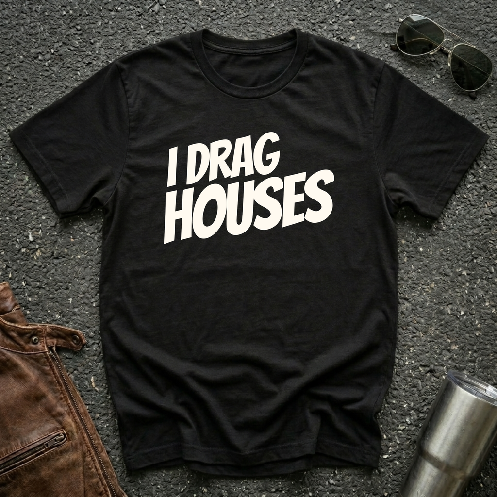 A black short-sleeve t-shirt with the phrase 'I DRAG HOUSES' printed in bold  letters across the chest, laid flat on a dark asphalt-like surface with sunglasses, brown leather jacket and a metal tumbler nearby.
