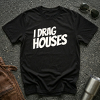A black short-sleeve t-shirt with the phrase 'I DRAG HOUSES' printed in bold  letters across the chest, laid flat on a dark asphalt-like surface with sunglasses, brown leather jacket and a metal tumbler nearby.