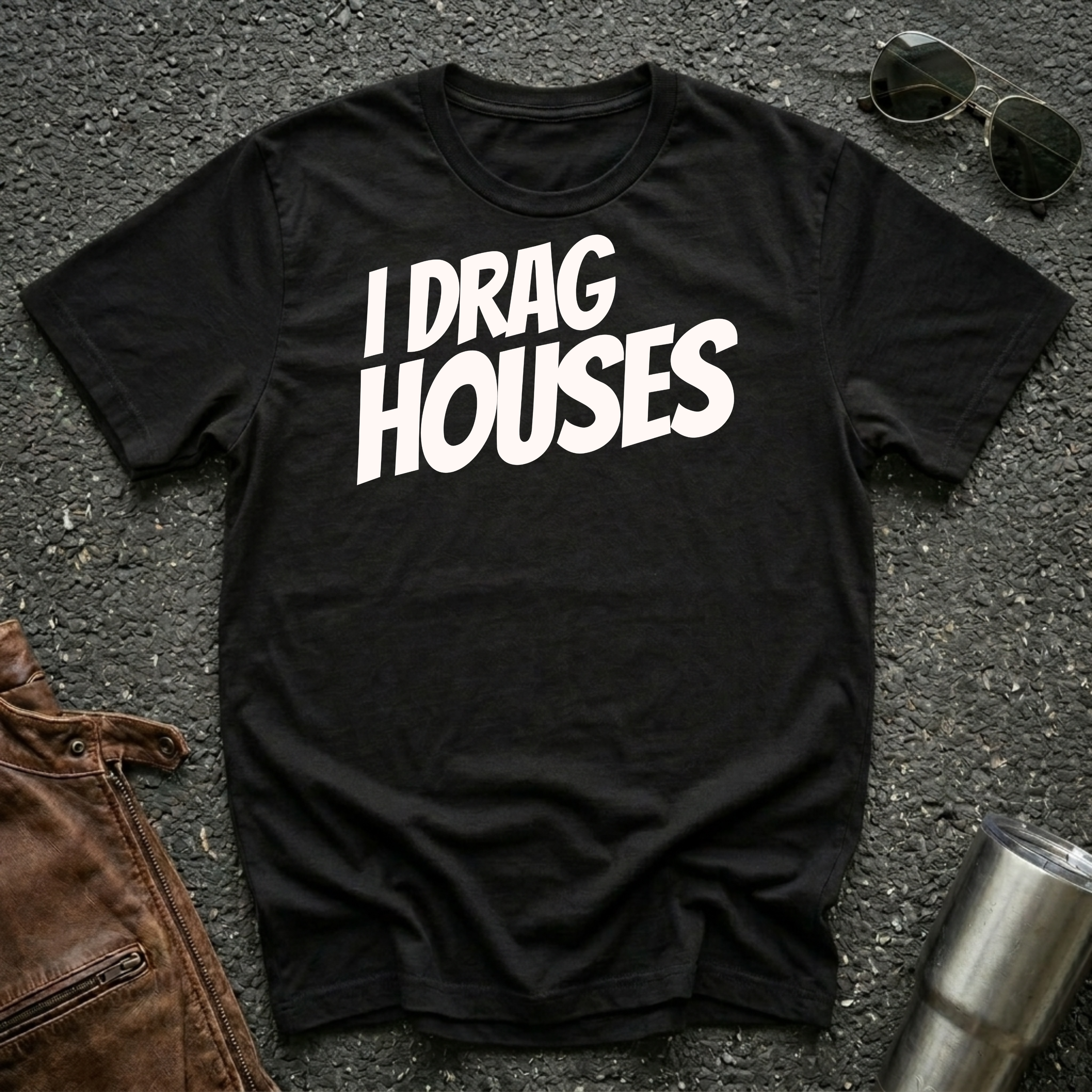 A black short-sleeve t-shirt with the phrase 'I DRAG HOUSES' printed in bold  letters across the chest, laid flat on a dark asphalt-like surface with sunglasses, brown leather jacket and a metal tumbler nearby.
