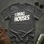 A dark heather short-sleeve t-shirt with the phrase 'I DRAG HOUSES' printed in bold  letters across the chest, laid flat on a dark asphalt-like surface with sunglasses, brown leather jacket and a metal tumbler nearby.