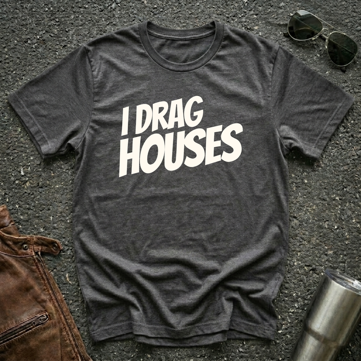 A dark heather short-sleeve t-shirt with the phrase 'I DRAG HOUSES' printed in bold  letters across the chest, laid flat on a dark asphalt-like surface with sunglasses, brown leather jacket and a metal tumbler nearby.