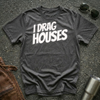 A dark heather short-sleeve t-shirt with the phrase 'I DRAG HOUSES' printed in bold  letters across the chest, laid flat on a dark asphalt-like surface with sunglasses, brown leather jacket and a metal tumbler nearby.