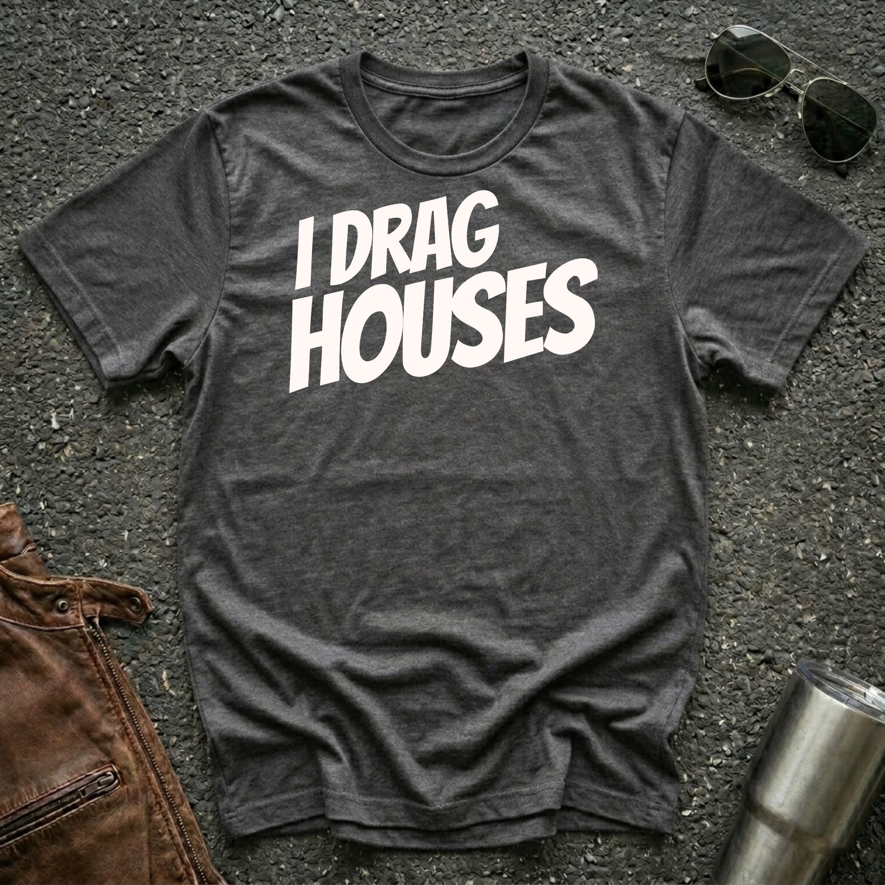 A dark heather short-sleeve t-shirt with the phrase 'I DRAG HOUSES' printed in bold  letters across the chest, laid flat on a dark asphalt-like surface with sunglasses, brown leather jacket and a metal tumbler nearby.