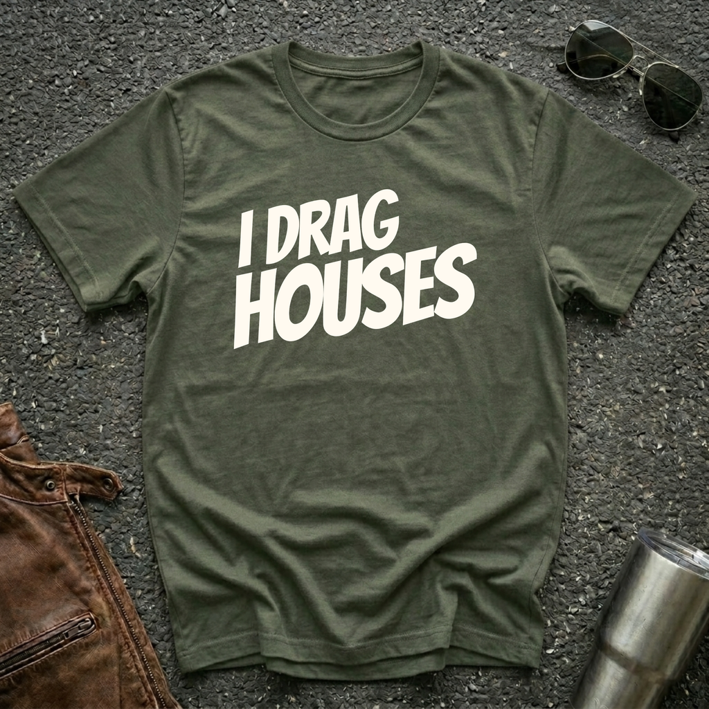 A military green short-sleeve t-shirt with the phrase 'I DRAG HOUSES' printed in bold  letters across the chest, laid flat on a dark asphalt-like surface with sunglasses, brown leather jacket and a metal tumbler nearby.
