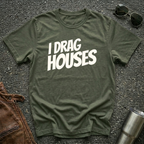 A military green short-sleeve t-shirt with the phrase 'I DRAG HOUSES' printed in bold  letters across the chest, laid flat on a dark asphalt-like surface with sunglasses, brown leather jacket and a metal tumbler nearby.