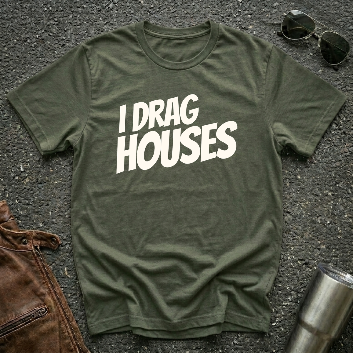 A military green short-sleeve t-shirt with the phrase 'I DRAG HOUSES' printed in bold  letters across the chest, laid flat on a dark asphalt-like surface with sunglasses, brown leather jacket and a metal tumbler nearby.