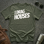 A military green short-sleeve t-shirt with the phrase 'I DRAG HOUSES' printed in bold  letters across the chest, laid flat on a dark asphalt-like surface with sunglasses, brown leather jacket and a metal tumbler nearby.