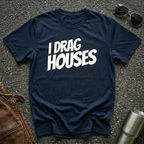 A navy short-sleeve t-shirt with the phrase 'I DRAG HOUSES' printed in bold  letters across the chest, laid flat on a dark asphalt-like surface with sunglasses, brown leather jacket and a metal tumbler nearby.