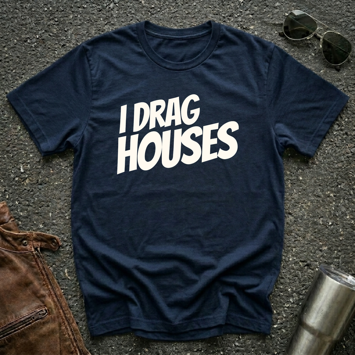 A navy short-sleeve t-shirt with the phrase 'I DRAG HOUSES' printed in bold  letters across the chest, laid flat on a dark asphalt-like surface with sunglasses, brown leather jacket and a metal tumbler nearby.