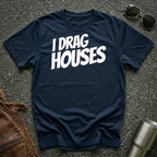 A navy short-sleeve t-shirt with the phrase 'I DRAG HOUSES' printed in bold  letters across the chest, laid flat on a dark asphalt-like surface with sunglasses, brown leather jacket and a metal tumbler nearby.